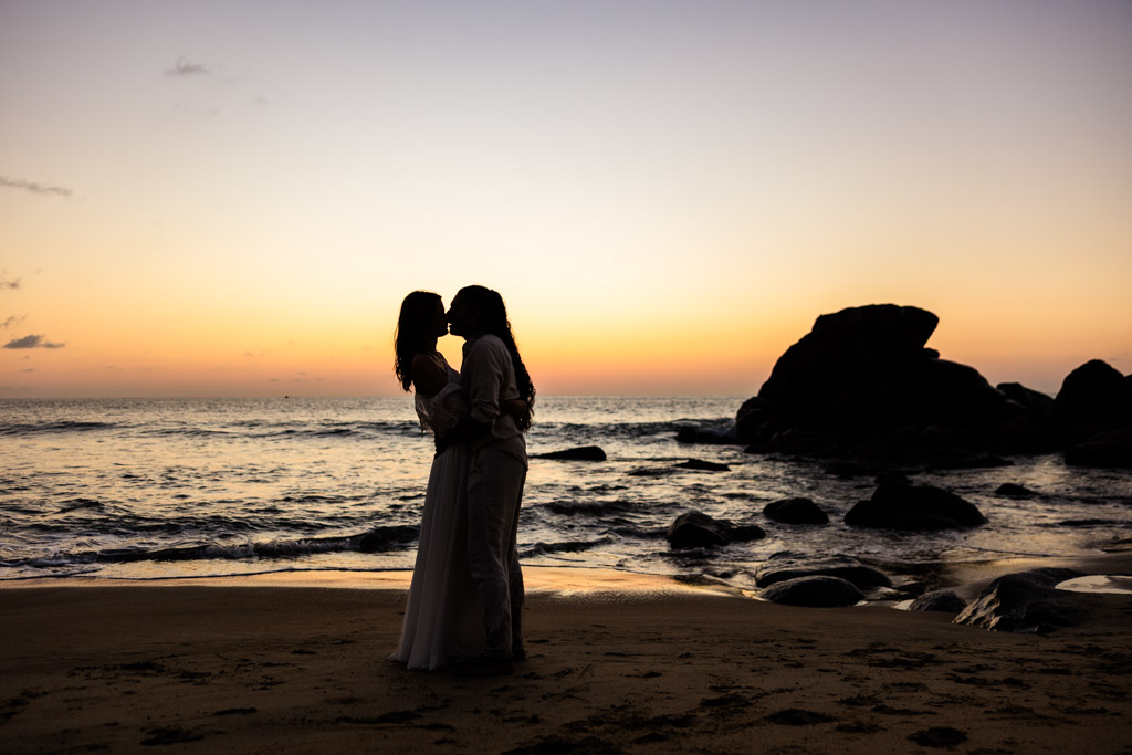 Two people embrace and kiss on a beach at sunset in Sayulita, silhouetted against the colorful sky and ocean during their engagement session