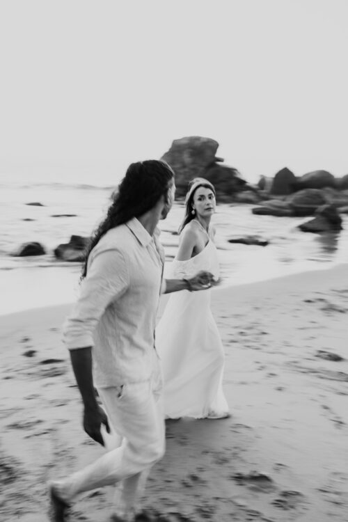 Creative black and white photo of couple in white clothes walking hand in hand along a beach, looking at each other during their Sayulita engagement session