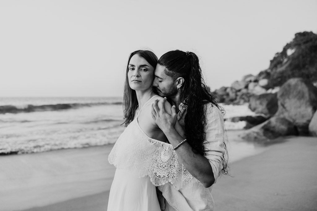 Black and white photo of couple on a Sayulita beach during their engagement session; the man kisses the woman's shoulder as she looks at the camera