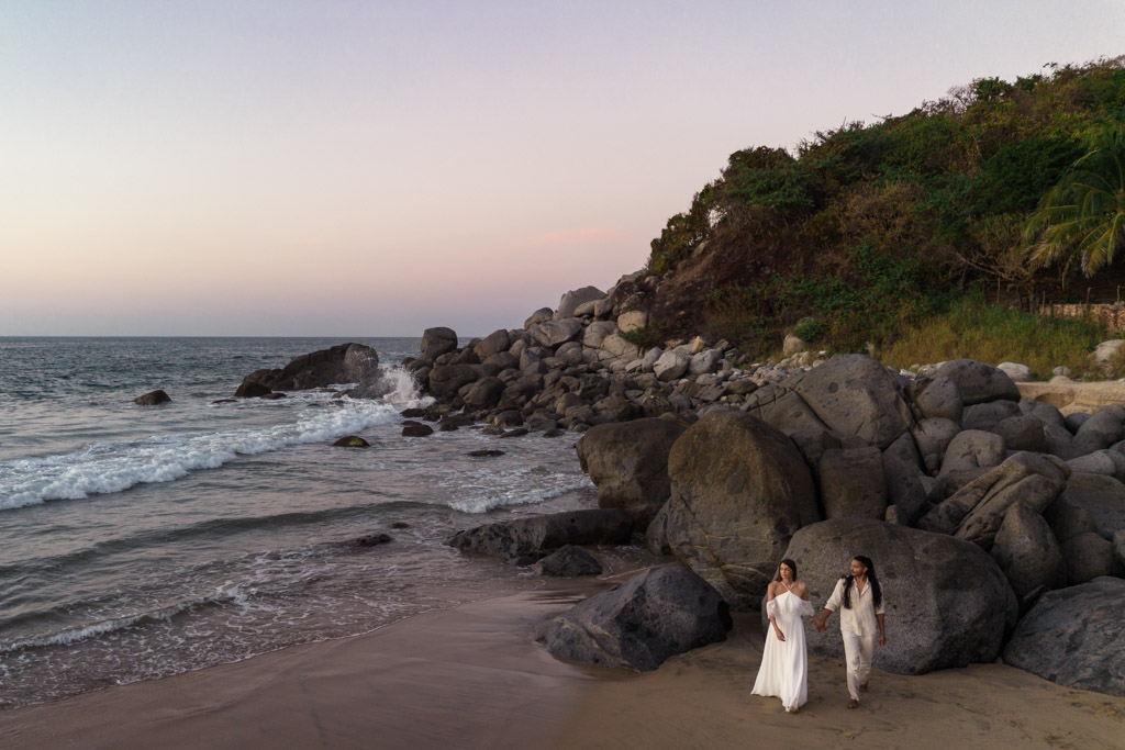Two people in wedding attire walk hand-in-hand on a rocky Sayulita beach at sunset with waves in the background