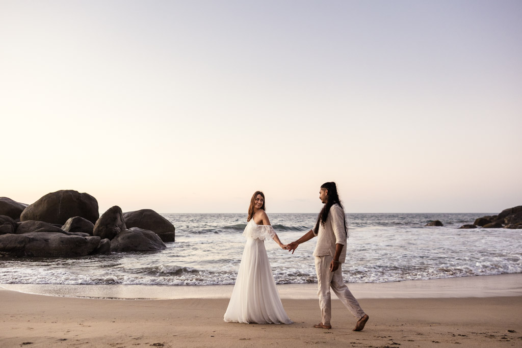 Couple holds hands and walks on a Sayulita beach at sunset, with rocks and the ocean in the background