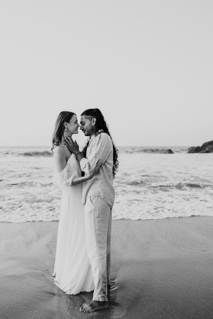 Black and white photo of couple standing close together on a beach during their Sayulita engagement session as waves crash in the background