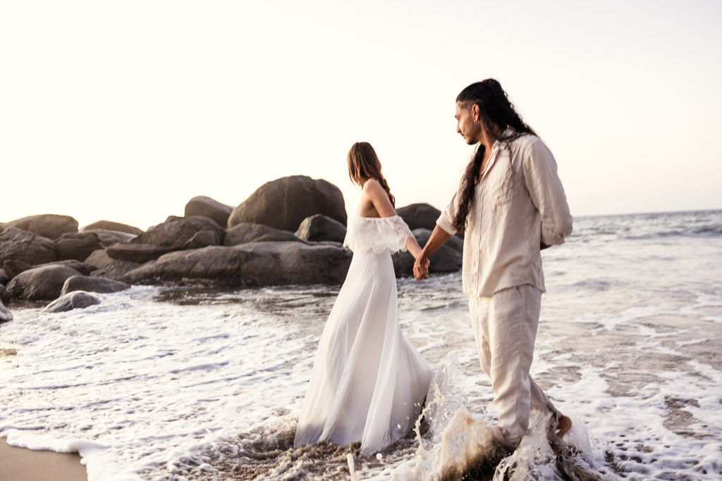 A couple holding hands walks along the beach shore during their Sayulita engagement session, waves splashing around them, with rocks in the background