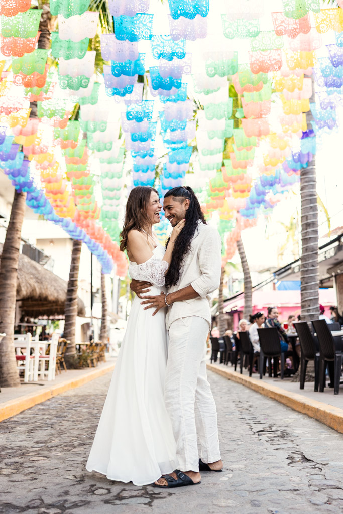 Happy couple in white stands on a cobblestone street under colorful hanging decorations, during their Sayulita engagement session