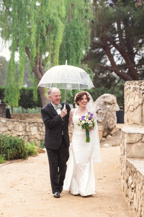 A bride in a white dress walks under a clear umbrella with an older man on a rainy day outdoors at a California vineyard wedding.