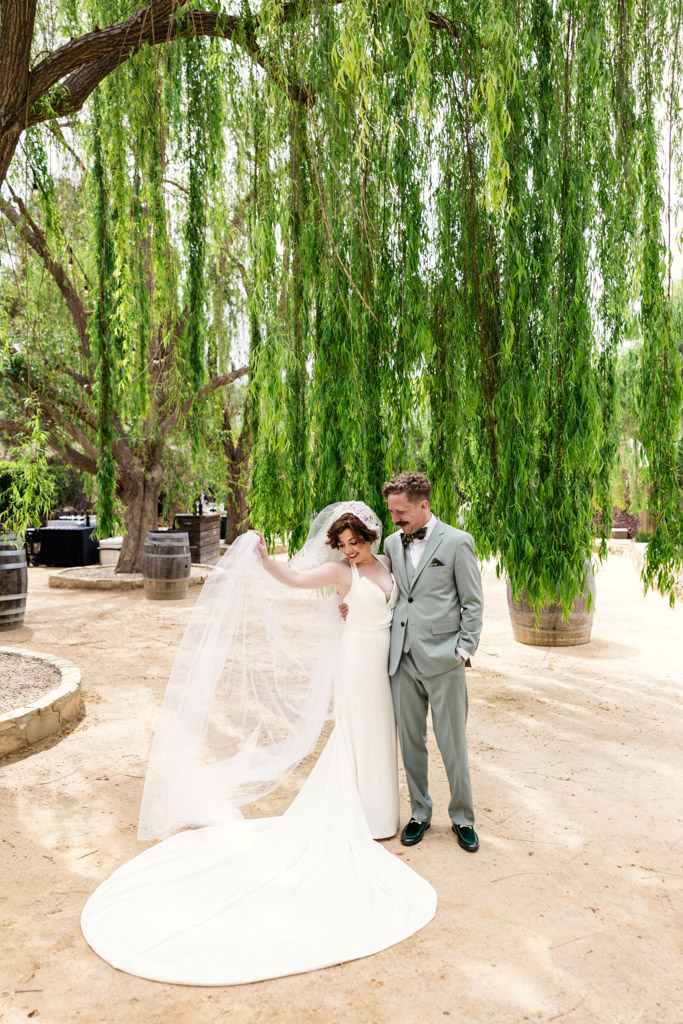 Bride and groom stand under willow trees at their Terra Mia Vineyard wedding celebration; bride holds out her long veil as the groom looks at her, smiling