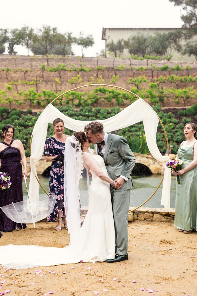 Bride and groom kiss under an arch outdoors, surrounded by smiling bridesmaids in dresses during their wedding ceremony at Terra Mia Vineyard