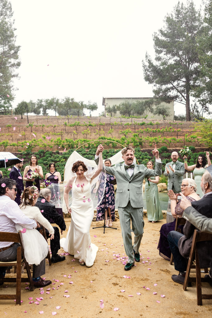 Bride and groom joyfully walk down the aisle as guests clap and toss flower petals after their wedding ceremony at Terra Mia Vineyard