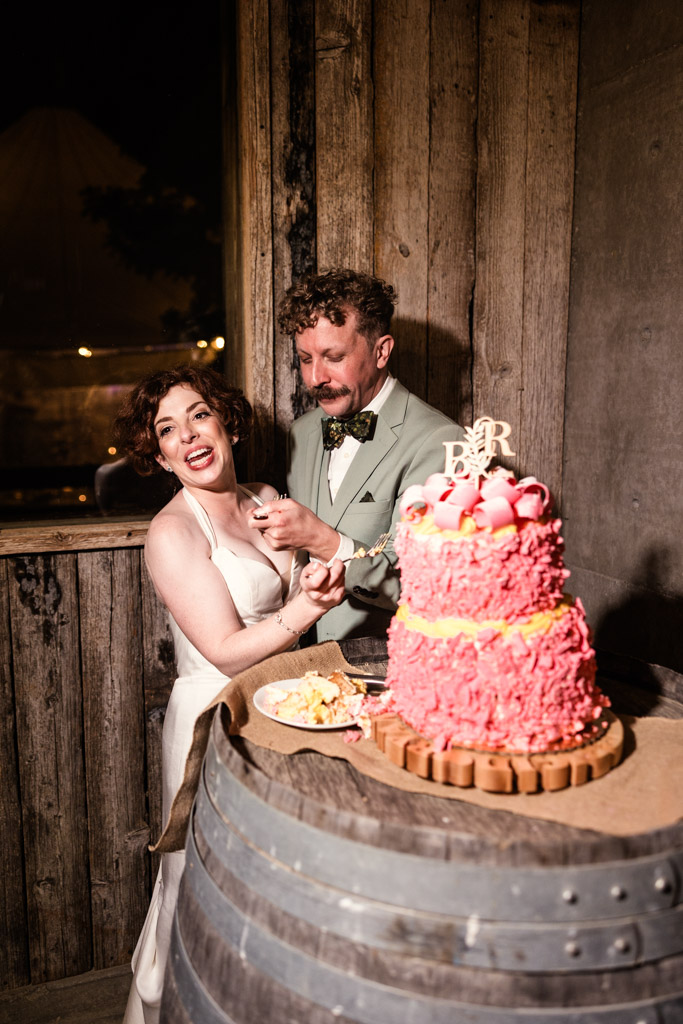Bride and groom laugh while eating their pink wedding cake together on a rustic barrel table at their Terra Mia Vineyard wedding reception