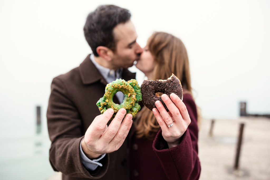 A couple kisses while holding donuts toward the camera on a foggy day by Lake Michigan during their Chicago winter engagement session