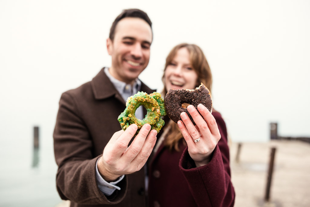 Happy engaged couple holding donuts close to the camera, standing outdoors by Lake Michigan during their Chicago winter engagement session