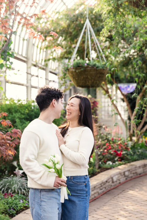 A couple smiles at each other in a bright greenhouse, holding flowers, surrounded by lush plants.