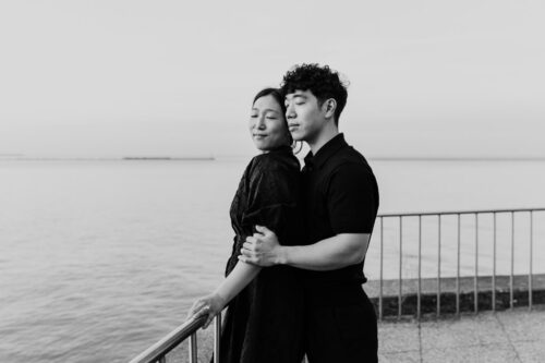 A couple embraces by a waterfront railing, both looking peaceful with eyes closed.
