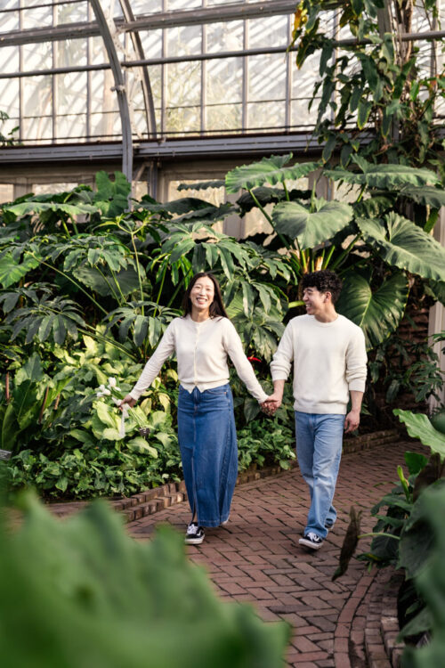A smiling couple holds hands and walks on a brick path in a lush greenhouse filled with large green plants.