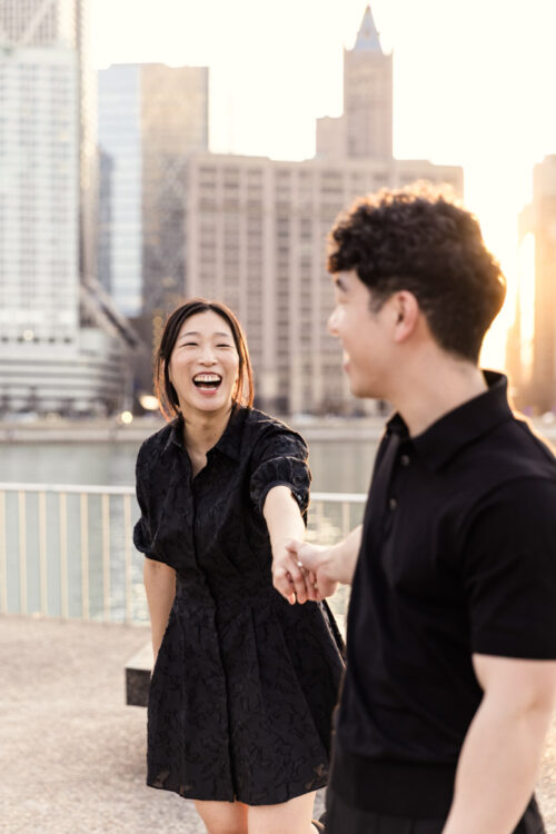 A woman in a black dress laughs while holding hands with a man in front of city buildings at sunset.