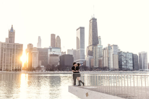 A couple embraces on a railing by the water with the Chicago skyline and sunset in the background.