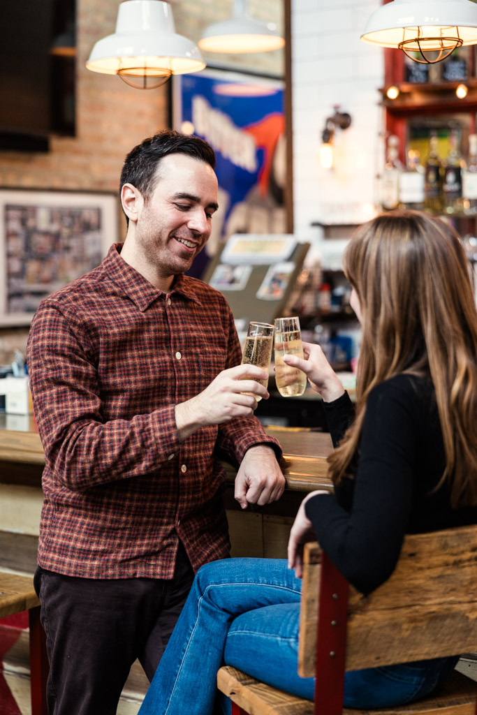 Two people sit at a bar, smiling and clinking glasses of champagne at Kibbitznest, during their Chicago winter engagement session