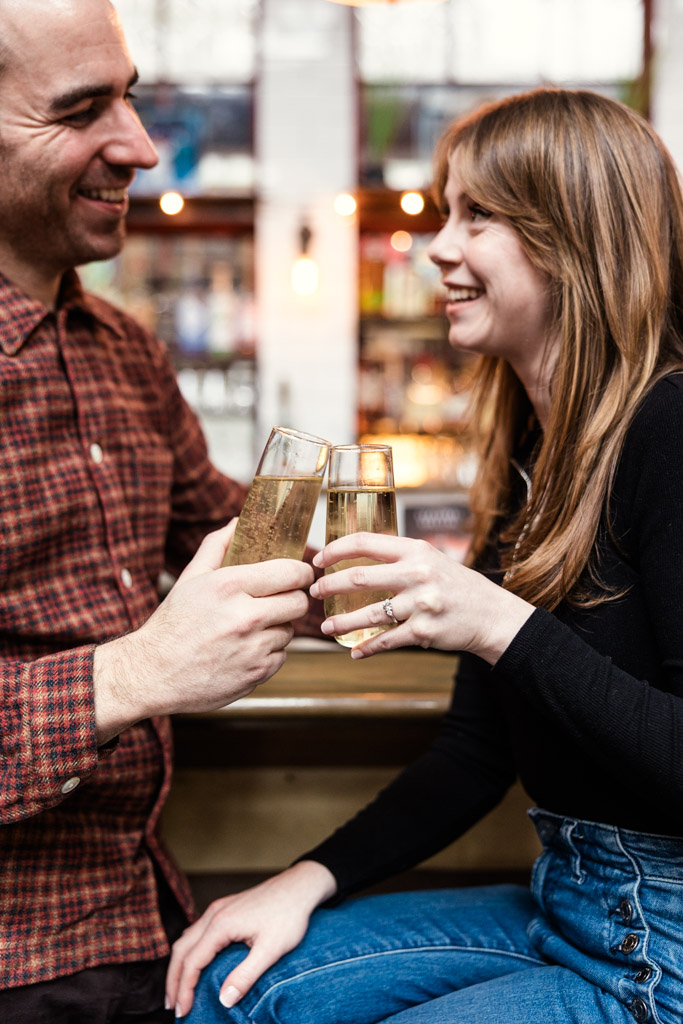 Two people smiling and clinking glasses of champagne together at a bar inside Kibbitznest during their Chicago winter engagement session