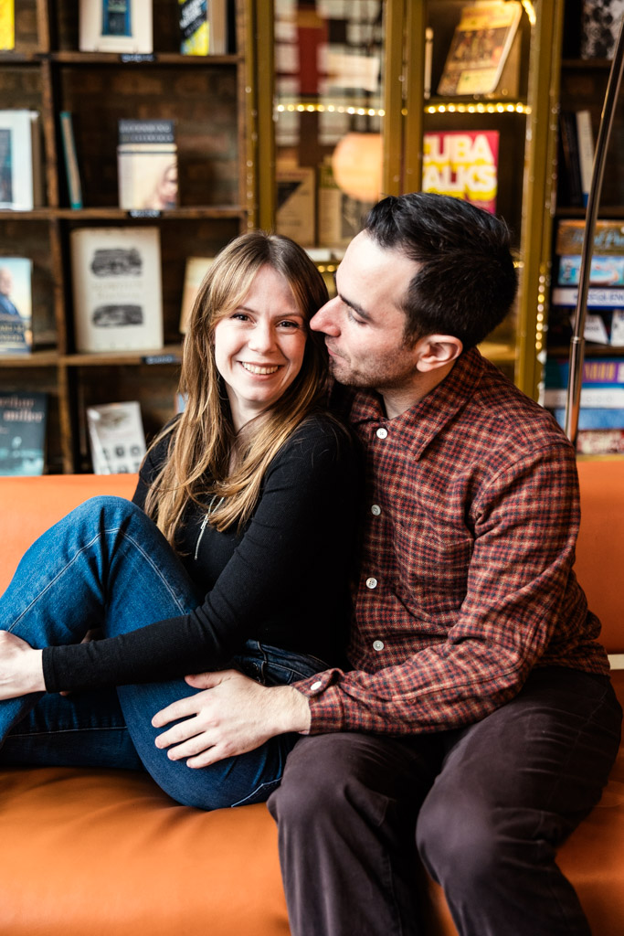 A smiling woman sits on an orange couch inside Kibbitznest beside her partner, with bookshelves in the background, during their Chicago winter engagement session