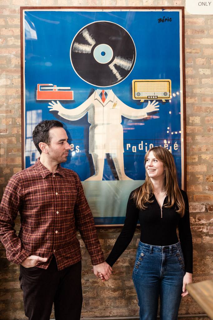 Engaged couple holding hands in front of a art poster featuring a figure with a vinyl record for a head inside Kibbitznest in Chicago