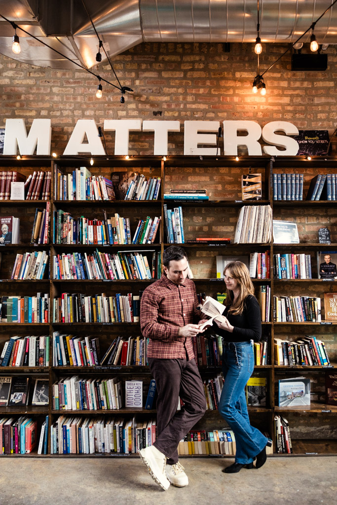Engaged couple reading books together in front of a large bookshelf at Kibbitznest