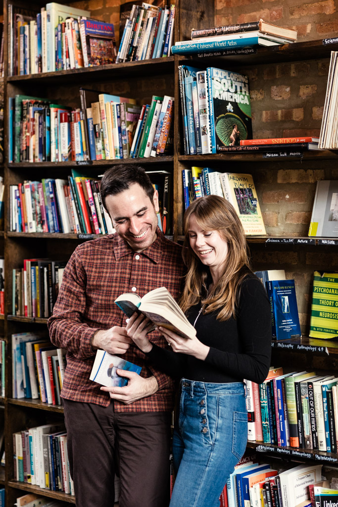 A man and woman stand inside Kibitznest, smiling and reading a book together in front of full bookshelves during their cozy Chicago winter engagement session