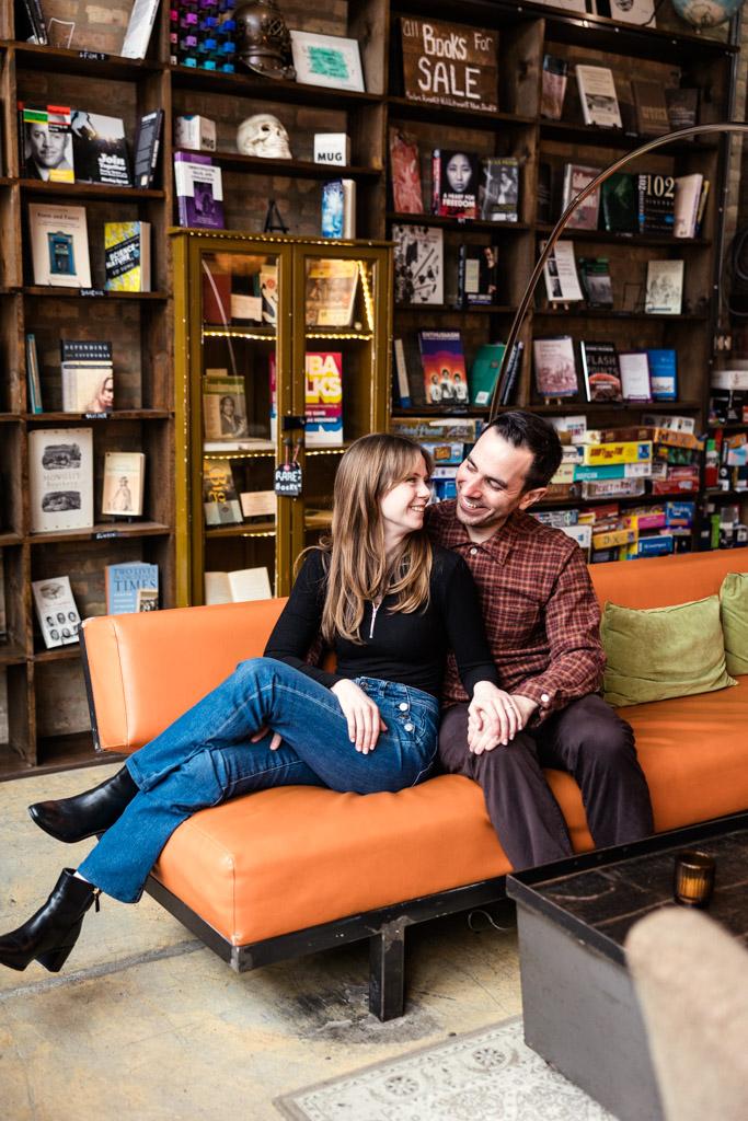 A smiling couple sits on an orange couch inside Kibbitznest bookstore in Chicago, surrounded by shelves of books during their Chicago winter engagement session