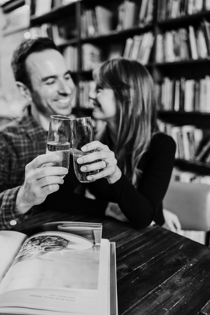 Black and white photo of happy engaged couple clinking glasses while sitting together inside Kibbitznest, with bookshelves in the background