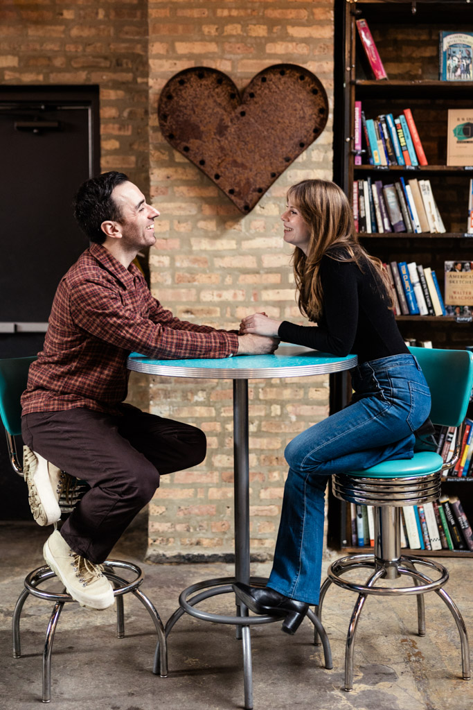 Engaged couple sits at a round table inside Kibbitznest smiling at each other and holding hands