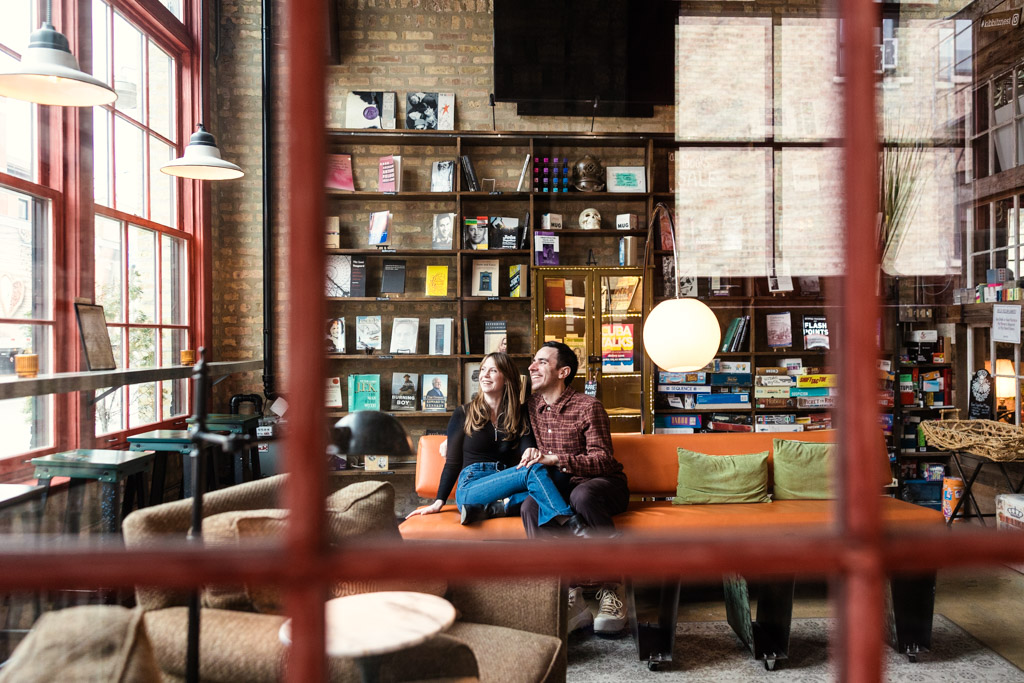 Engaged couple sits on an orange couch, inside Kibbitznest bookstore in Chicago with large windows and warm lighting