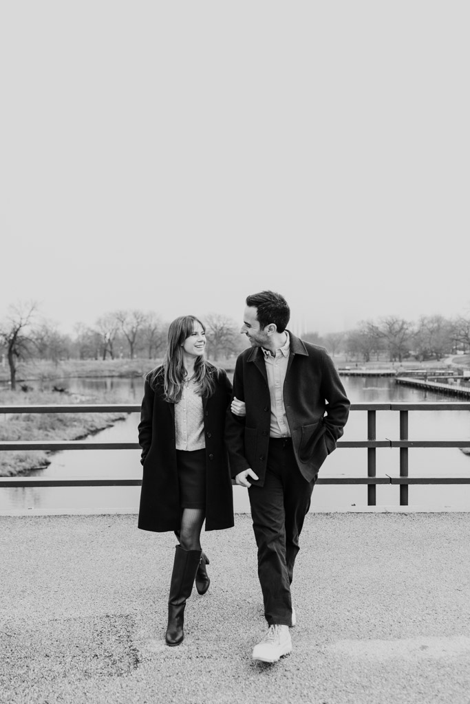 Black and white photo of couple walking arm-in-arm outdoors on a Lincoln Park bridge, smiling at each other during their Chicago winter engagement session