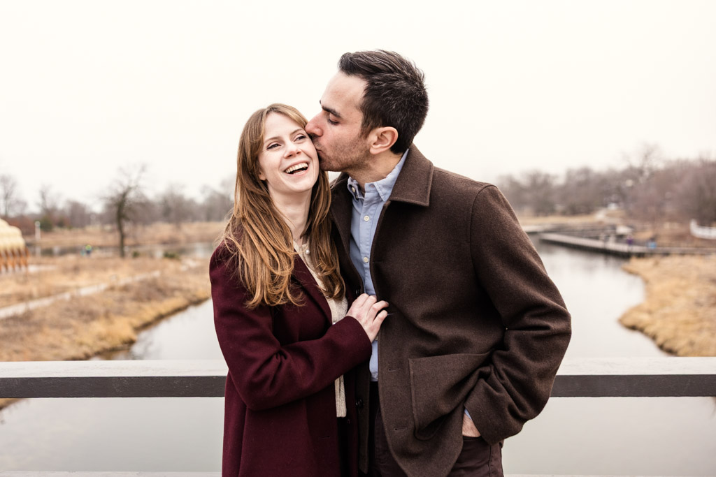 A man kisses a smiling woman on the cheek as they stand together on a Lincoln Park bridge over a river during their Chicago winter engagement session