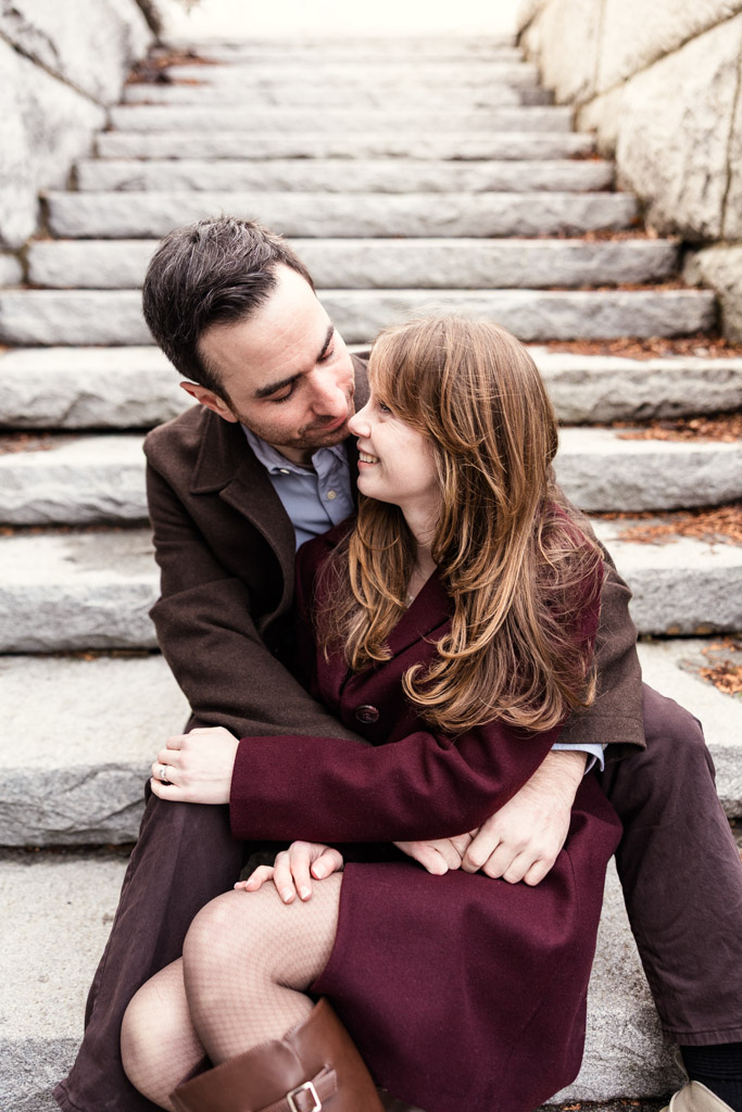 A couple in coats sits closely on stone steps, smiling and looking lovingly at each other during their Chicago winter engagement session