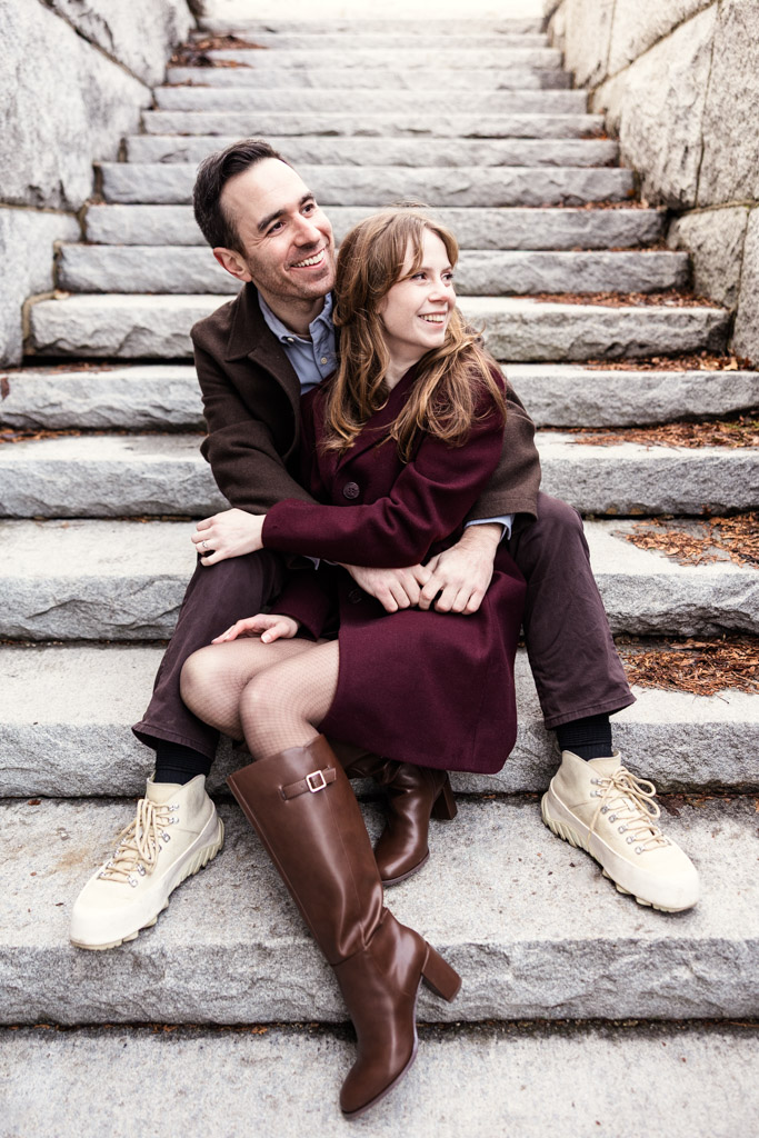 A smiling couple sits on stone steps, embracing and looking to the side during their Chicago winter engagement session