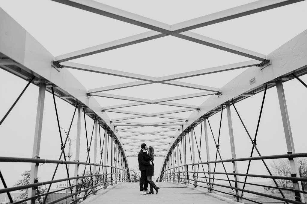Black and white photo of engaged couple on a Lincoln Park pedestrian bridge, framed by metal beams