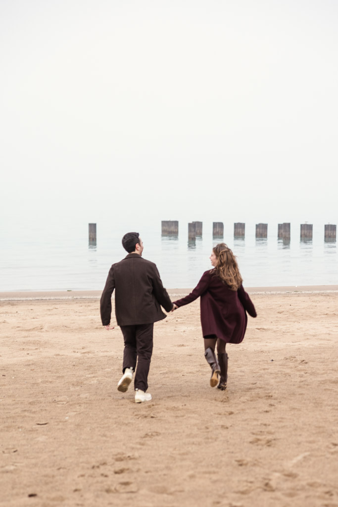 A couple holding hands runs on a sandy beach toward the water on a foggy day during their Chicago winter engagement session