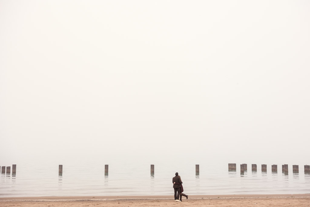 Engaged couple stands on a foggy Lake Michigan beach, facing calm water with wooden posts emerging from the lake