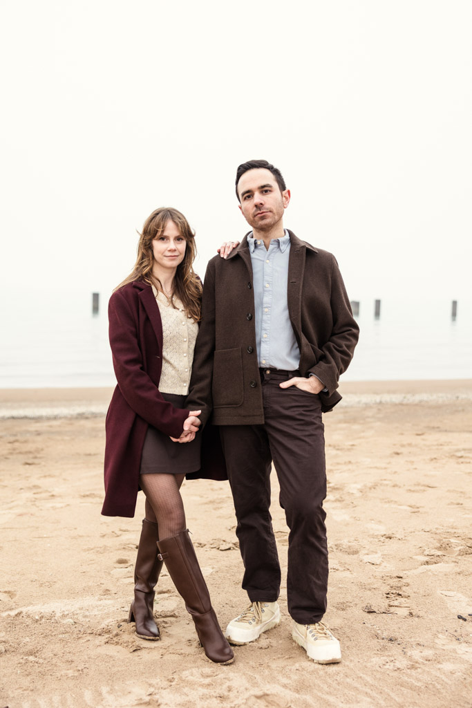 Portrait of couple on a sandy beach, holding hands and wearing dark coats, with calm water in the background during their foggy Chicago winter engagement session