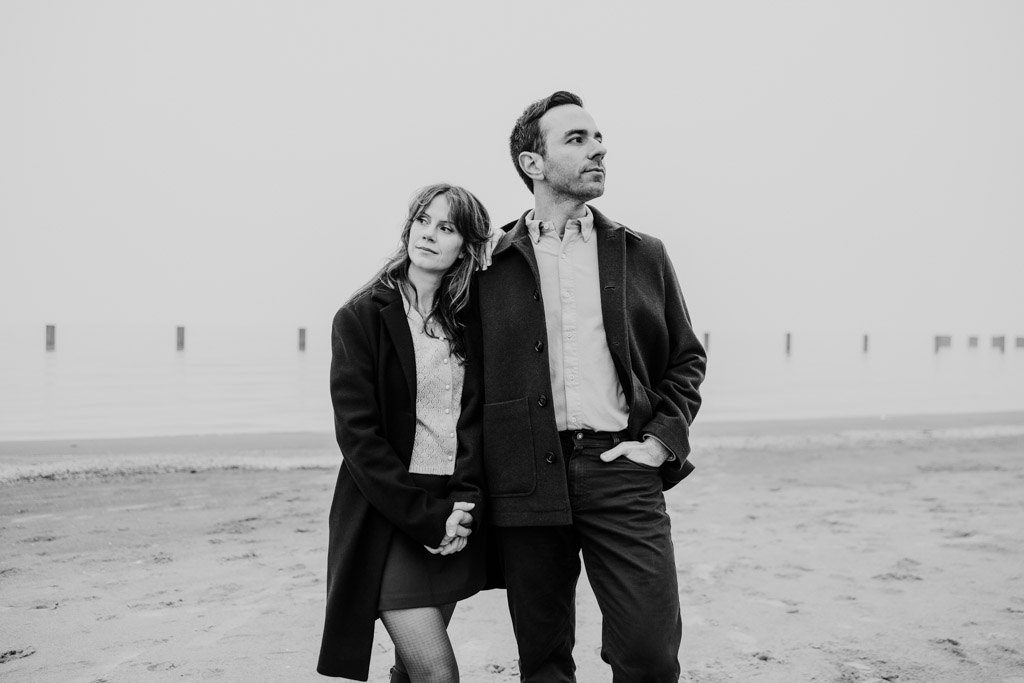 Blacka and white photo of couple standing on a Lake Michigan beach, holding hands and looking thoughtfully into the distance during their Chicago winter engagement session