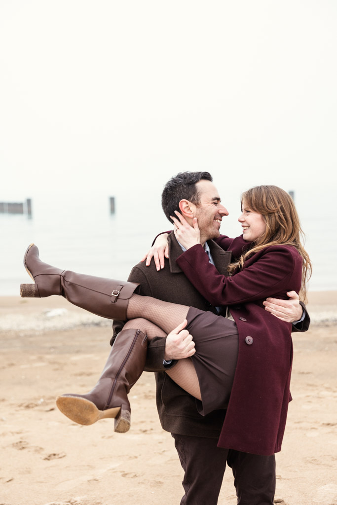 Man lifts and smiles at his aprtner on a sandy Lake Michigan beach during their Chicago winter engagement session