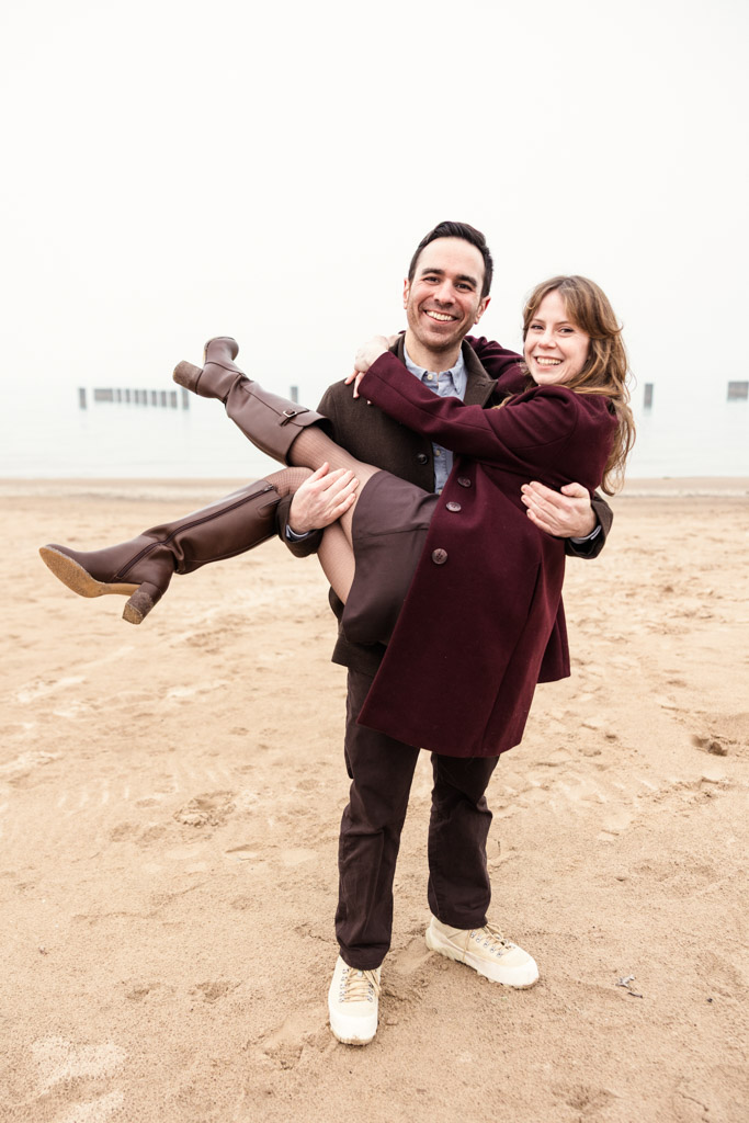 A man smiles while carrying his partner in his arms on a sandy Lake Michigan beach with foggy water in the background during their Chicago winter engagement session