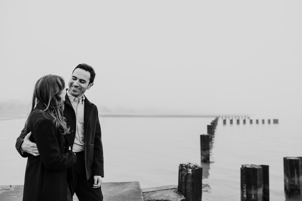 A couple stands close together, smiling by a foggy lakeshore with wooden posts in the water, during their Chicago winter engagement session