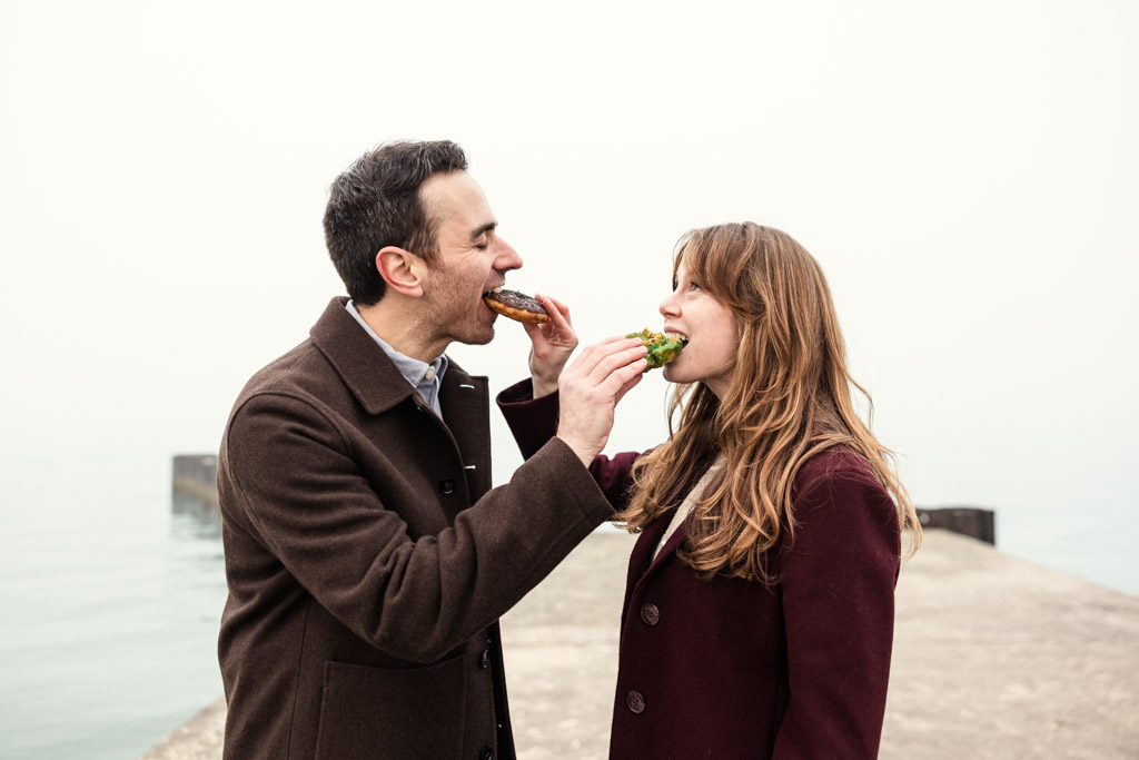 A man and woman smiling and feeding each other donuts on a foggy lakefront pier during their Chicago winter engagement session