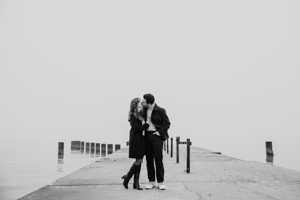 Black and white photo of engaged couple standing on a foggy pier, embracing and kissing during their Chicago winter engagement session, with calm water in the background.