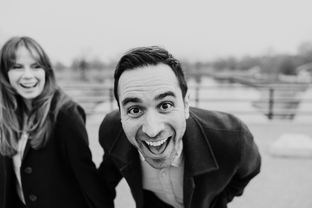Black and white photo of man smiling excitedly at the camera while his partner laughs beside him during their Chicago winter engagement session