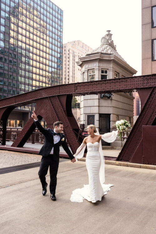 Bride and groom holding hands and celebrating on a city bridge, surrounded by tall buildings.