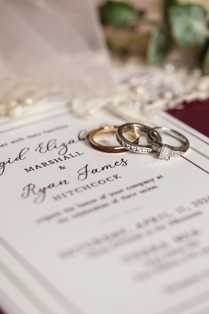 Three wedding rings rest on a wedding invitation with elegant script, set against blurred greenery for Terra Mia Vineyard wedding