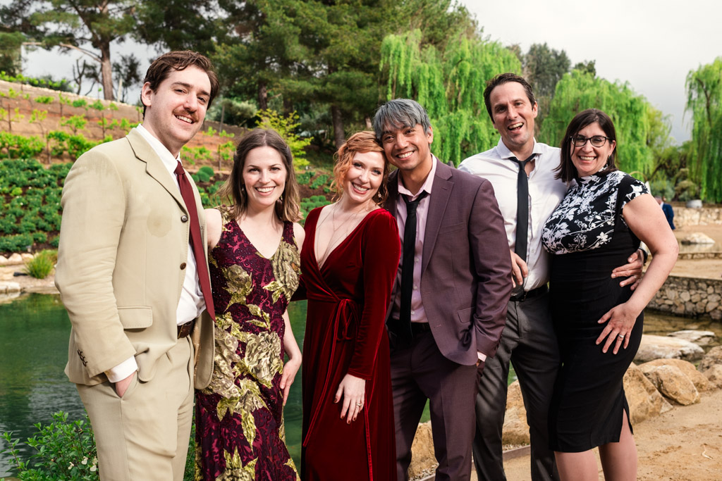 Six guests in formal attire stand smiling together outdoors by a pond during wedding celebration at Terra Mia Vineyard