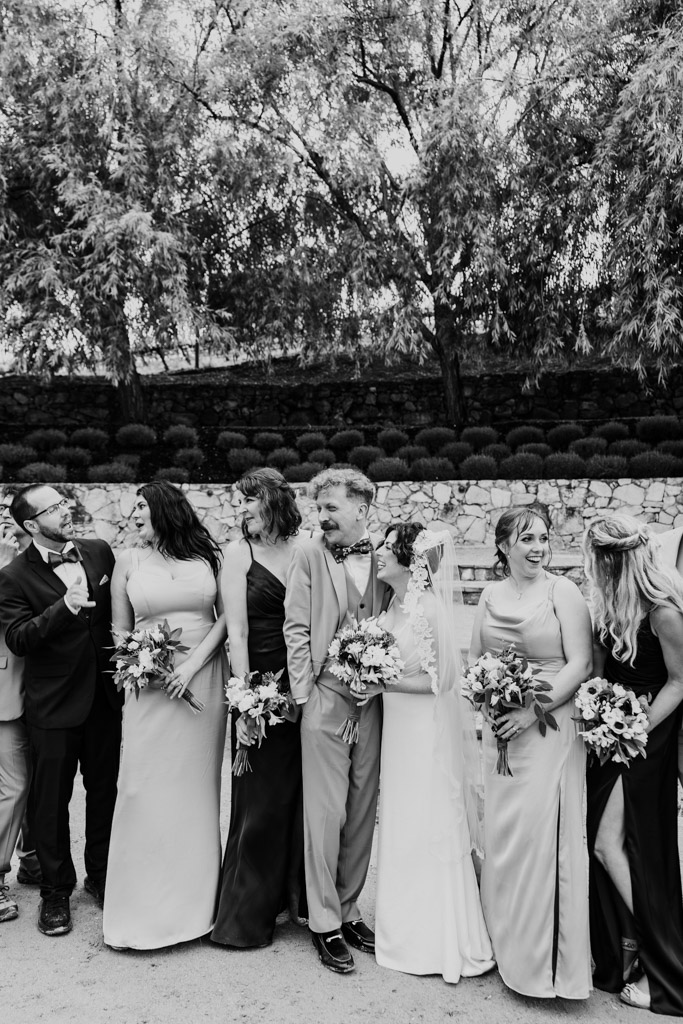 Black and white photo of wedding party outdoors, smiling and holding bouquets, with trees and stone walls in the background at Terra Mia Vineyard