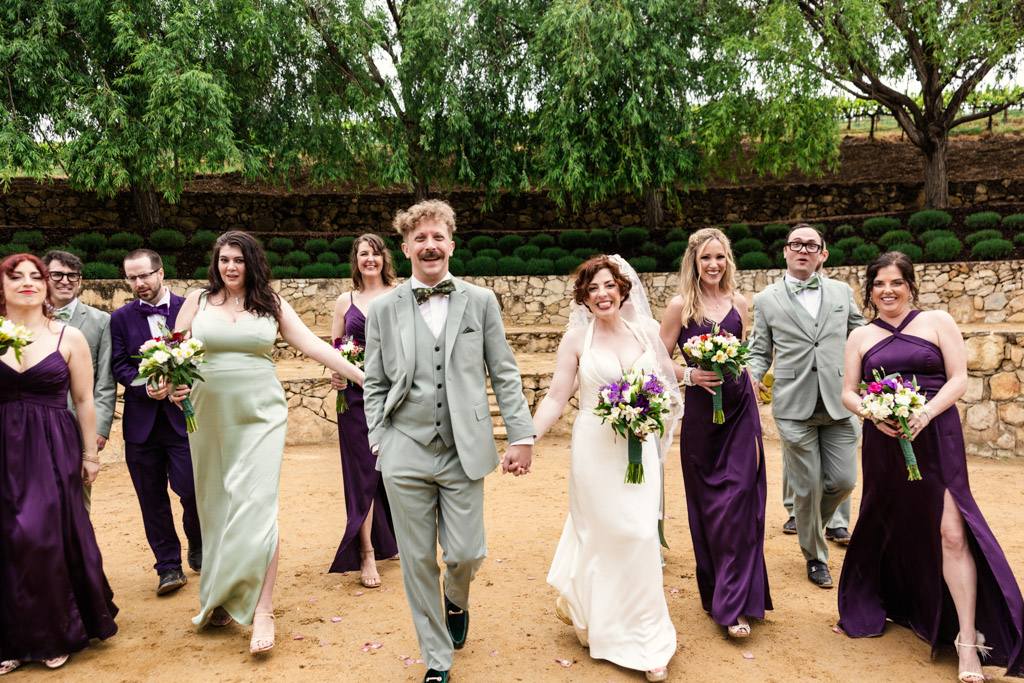 A wedding party smiles and walks outdoors at Terra Mia Vineyard wedding celebration, with the bride and groom holding hands in the center